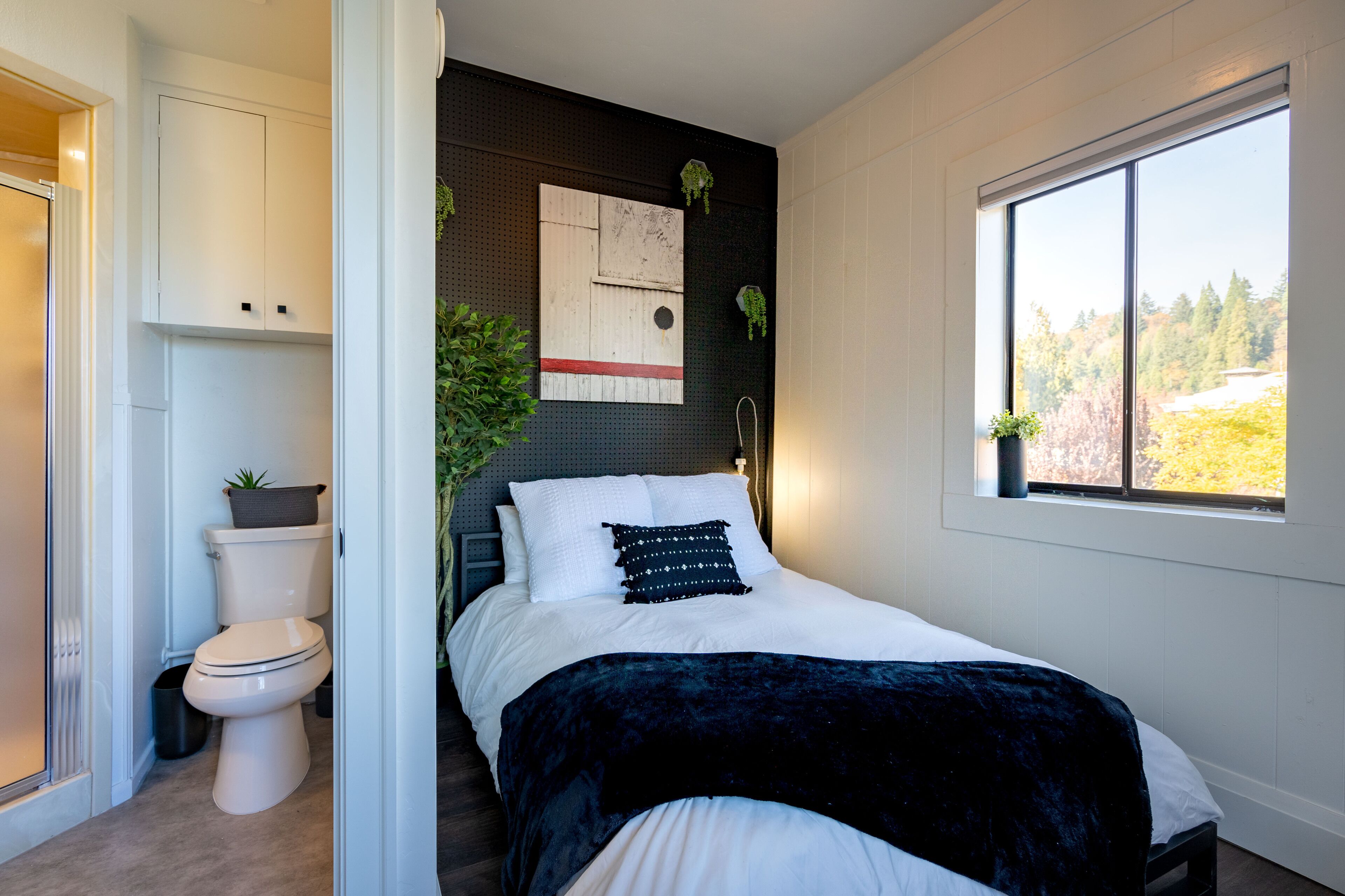 A bedroom with a bed, a window, a wall-mounted artwork, and a view of trees outside.