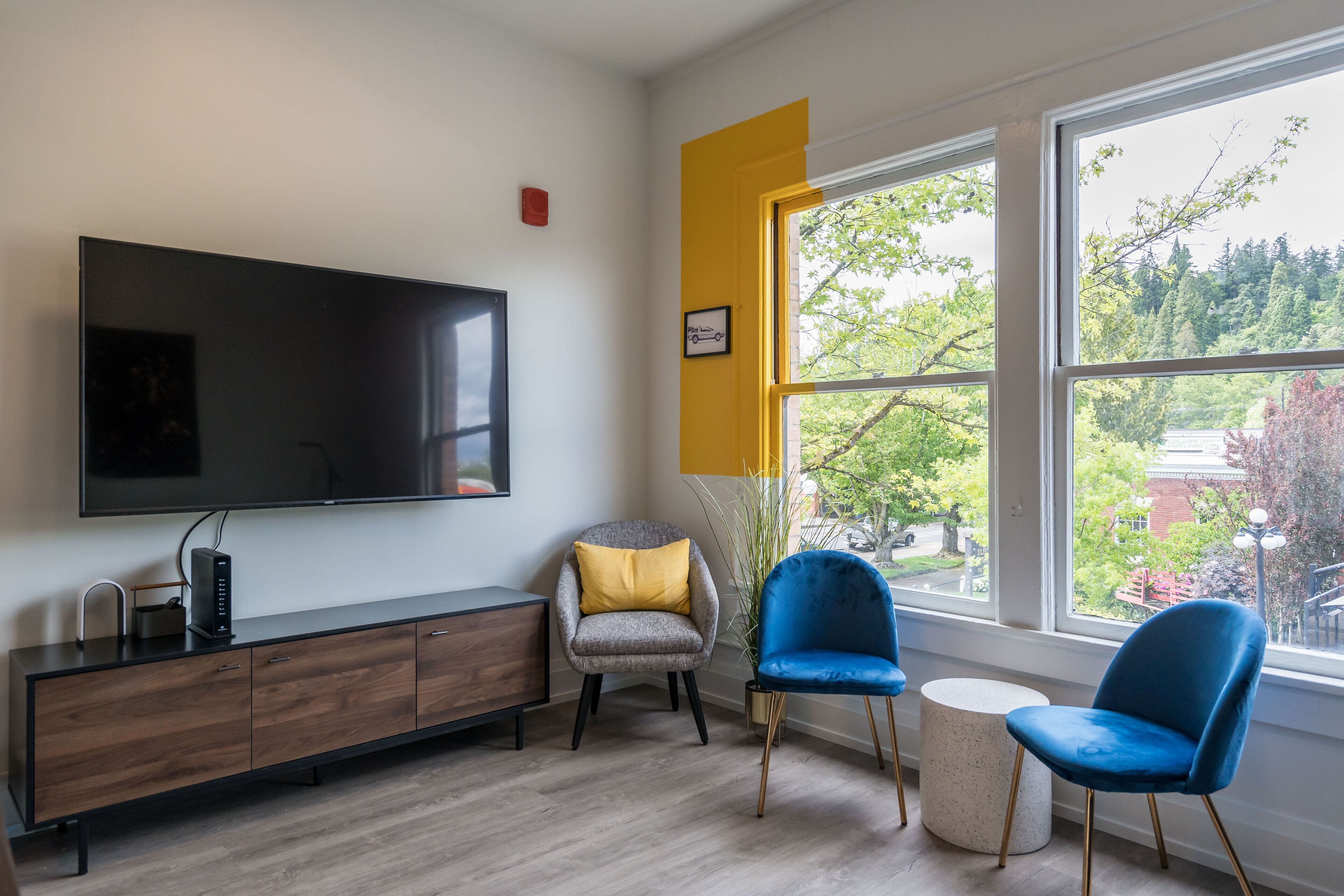 Un salon moderne doté d’un grand téléviseur à écran plat, d’un meuble en bois, de deux fauteuils et d’une grande fenêtre donnant sur des arbres et des immeubles.