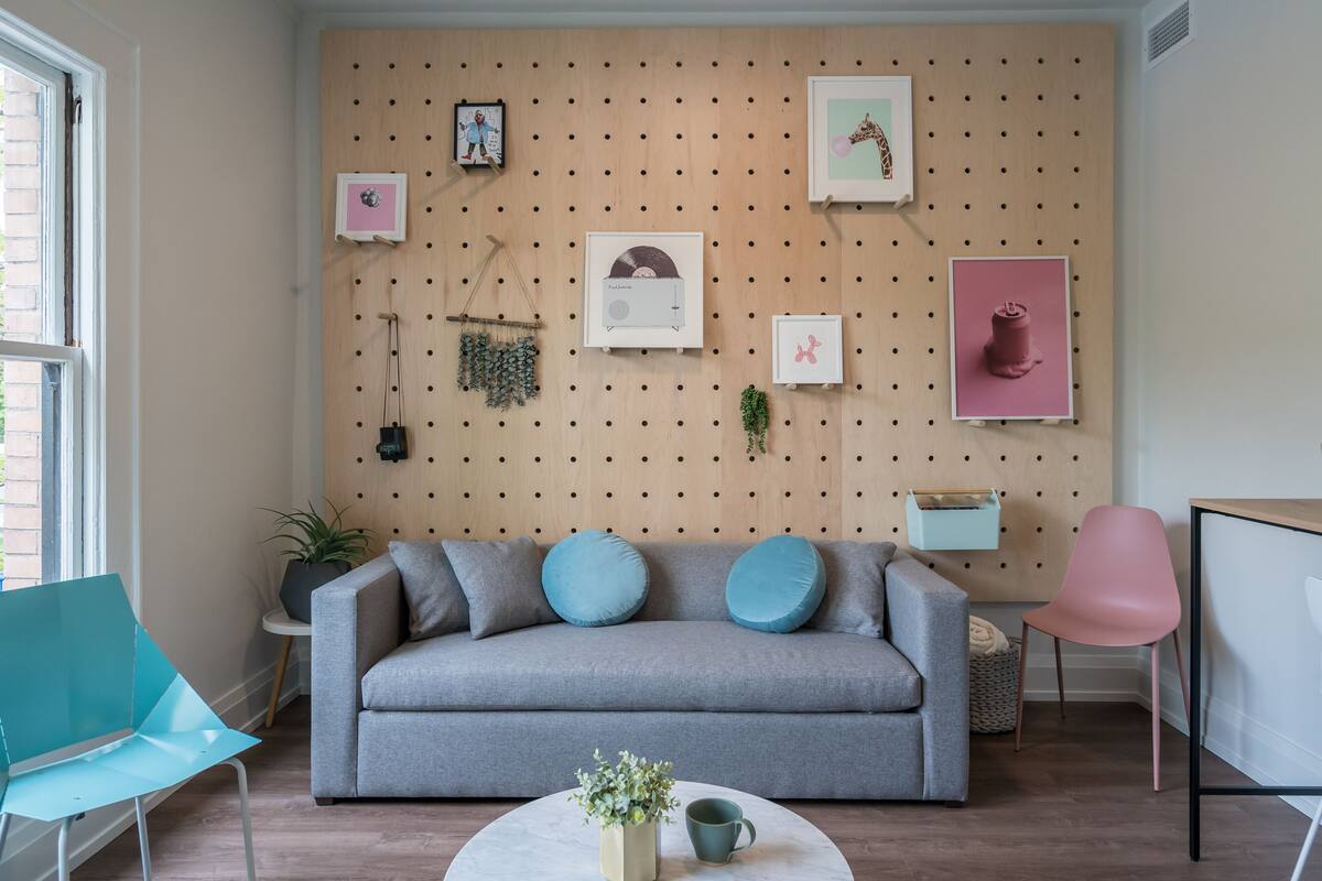 A modern living room with a grey sofa, teal and blue cushions, a round coffee table with a plant and mug, and a wooden wall with various framed pictures and a hanging plant.