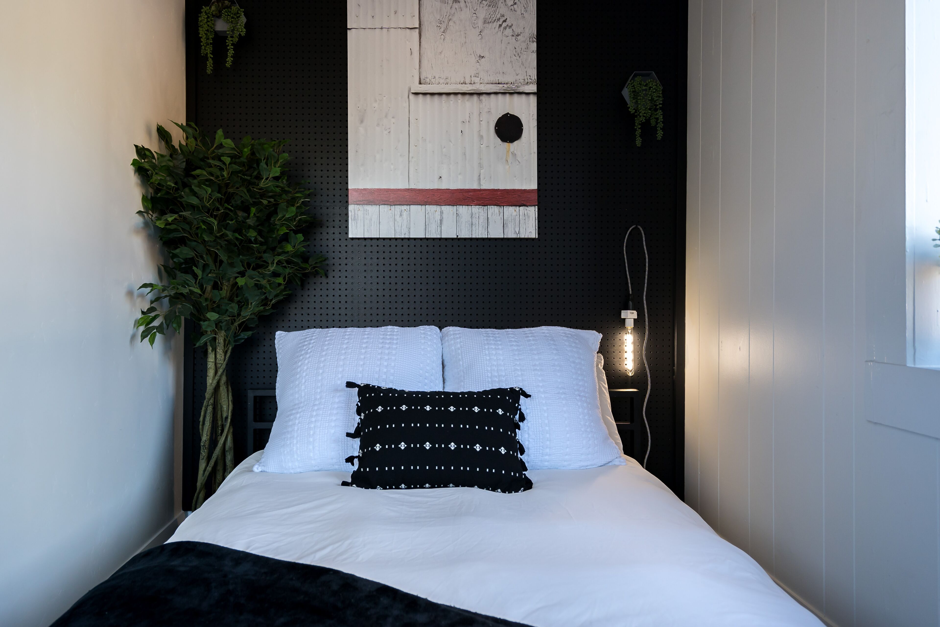 Una cama bien tendida con ropa blanca y almohadones decorativos, una lámpara de pared y una planta en maceta.