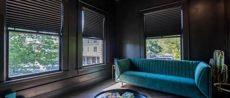 Uma sala de estar moderna com um sofá verde-água, grandes janelas com persianas, uma mesa de centro redonda com livros e uma planta de cacto.