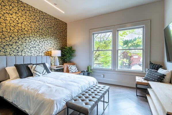A modern bedroom with a large bed, a bench, a TV, and a window with a view of trees. - The 505 Eugene (Eugene)