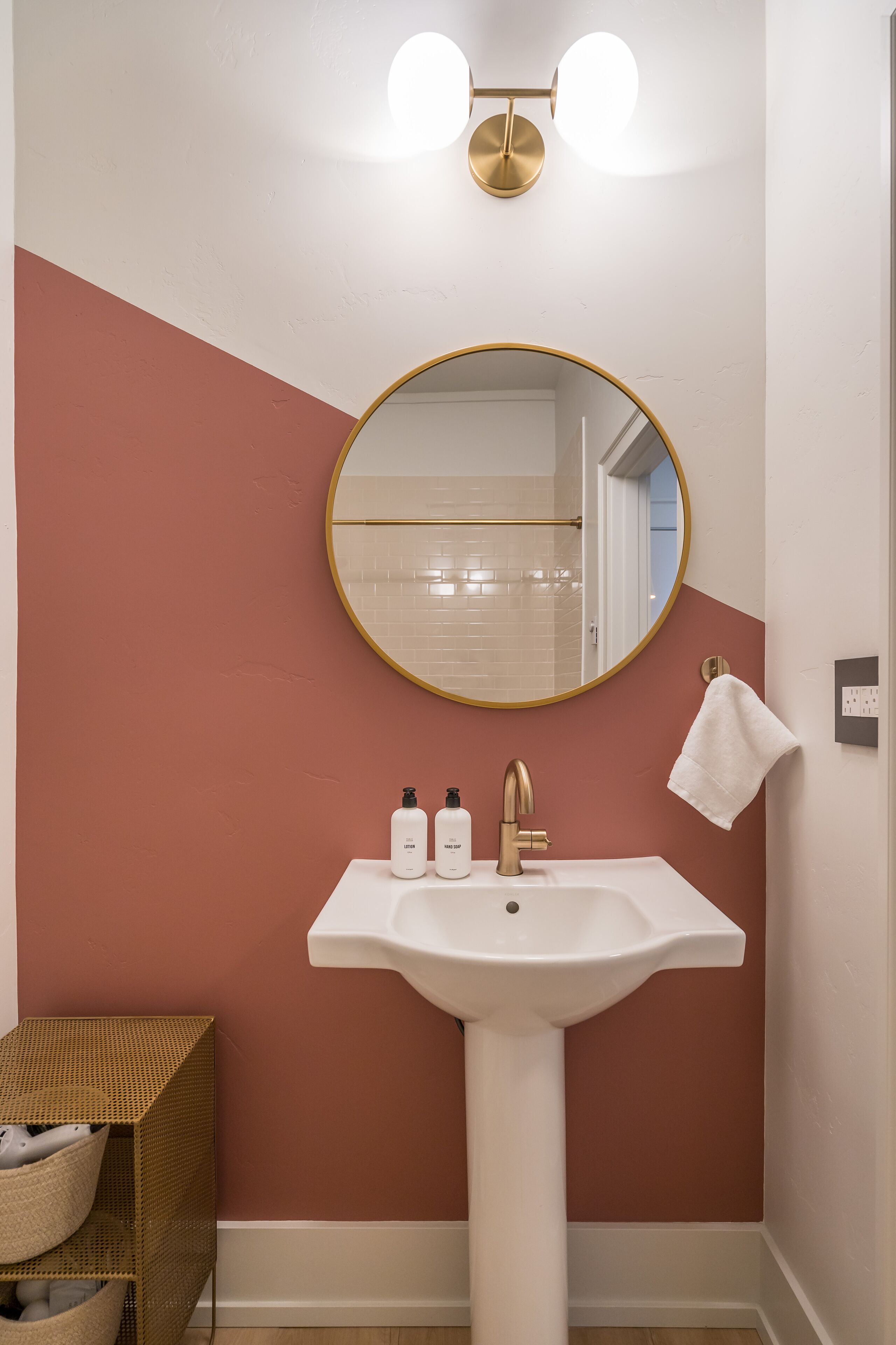 A bathroom with a white sink, gold faucet, and a round mirror with a gold frame. There are two bottles on the sink and a towel hanging on the wall.