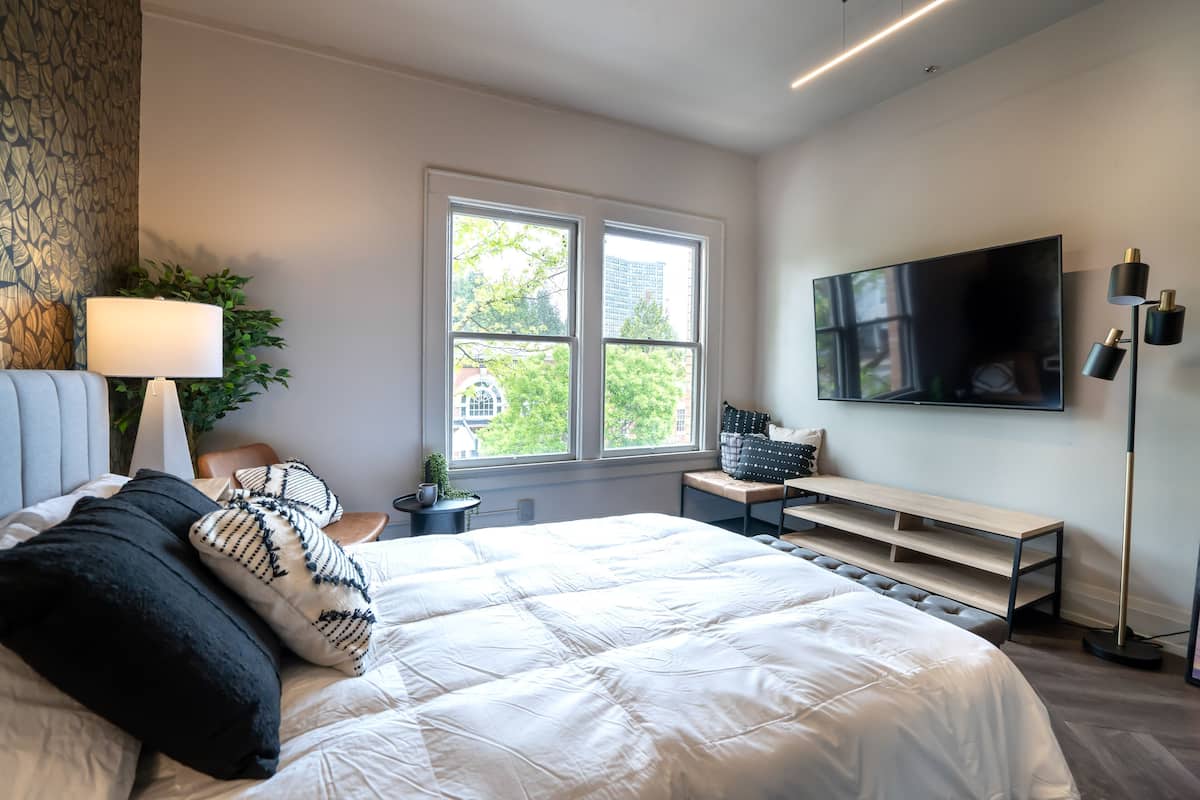 A modern bedroom with a large bed, a TV, a desk, and a window with a view of buildings and trees.