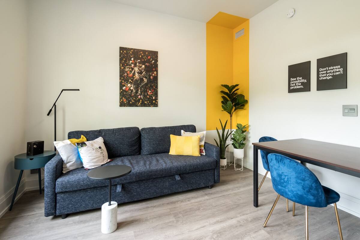 A modern living room with a grey sofa, a small round table, a dining table with blue chairs, a floor lamp, and a large plant.