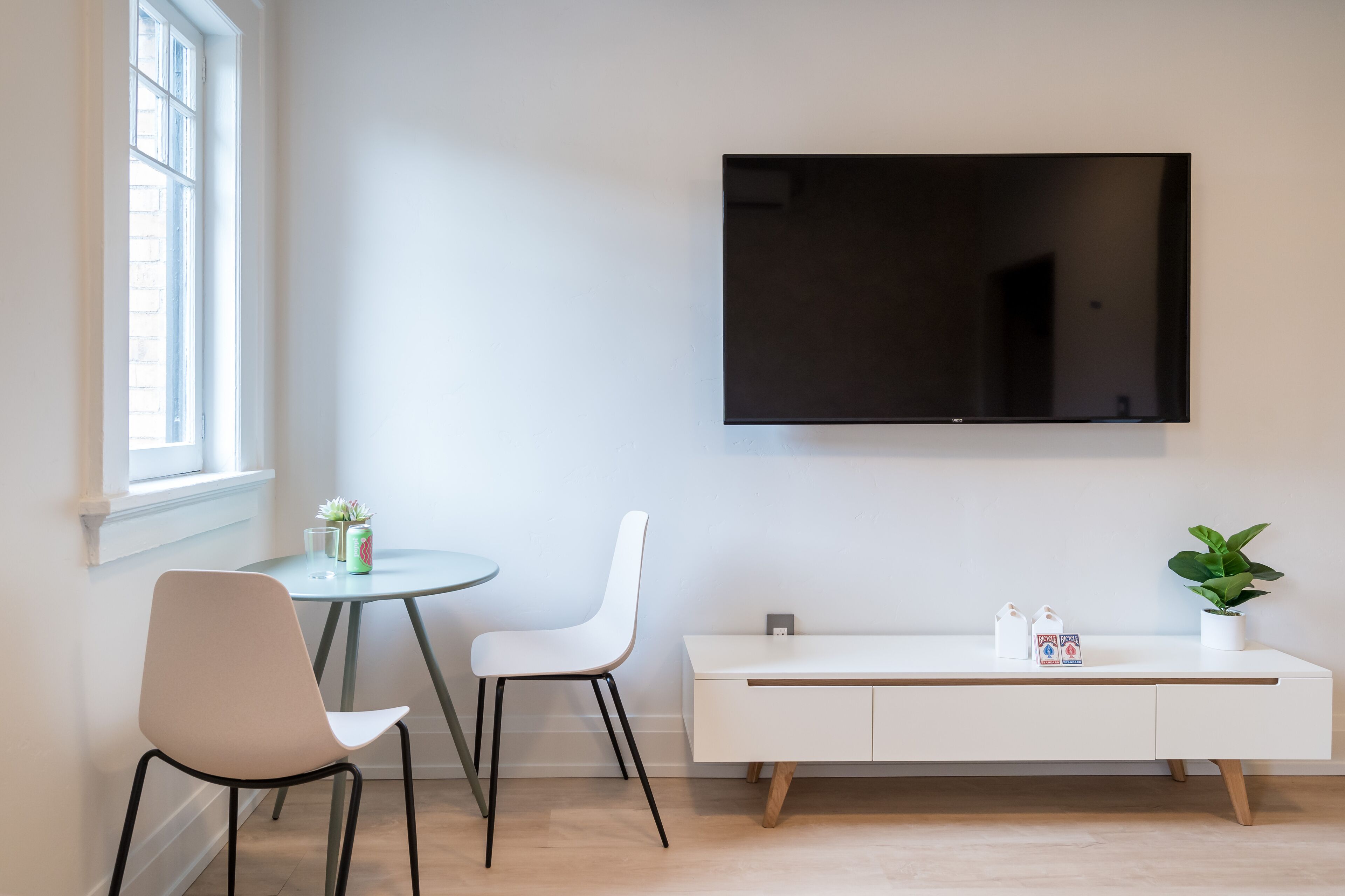 A modern living room with a flat-screen TV mounted on the wall, a white console table, two white chairs, a small round table with a potted plant, and a window providing natural light.