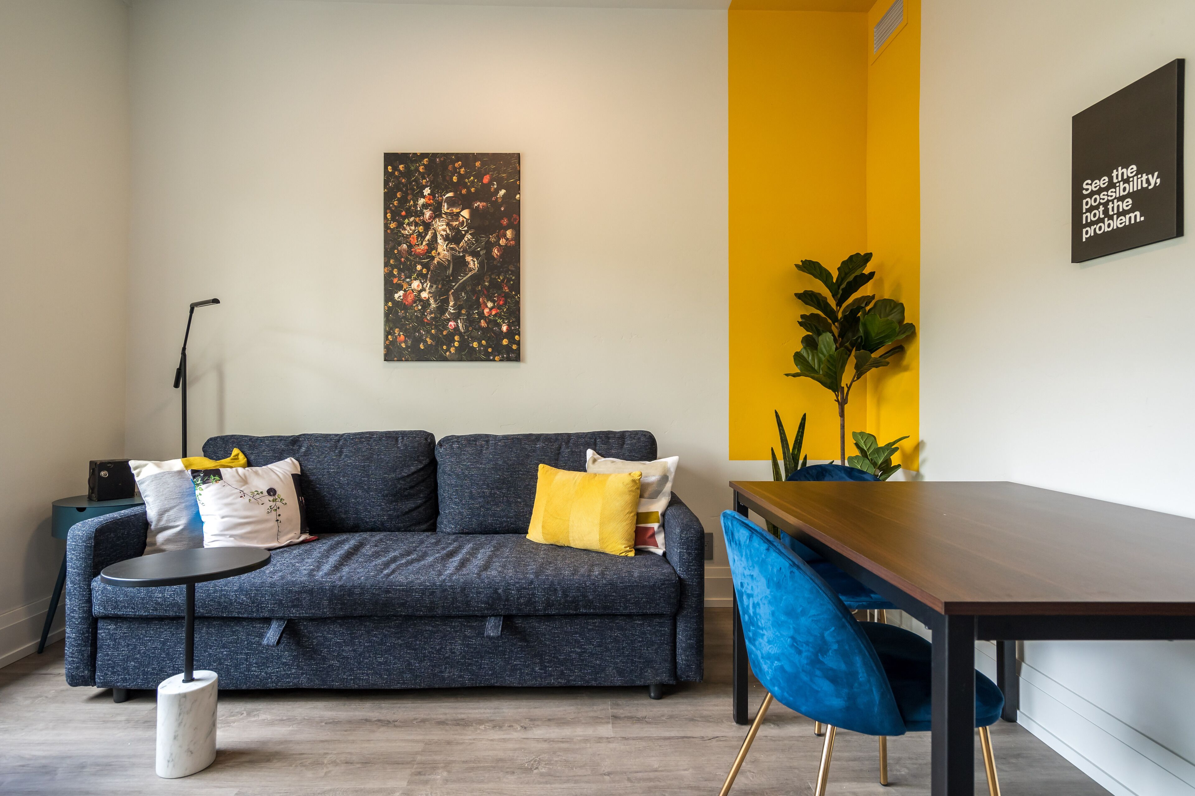 Uma sala de estar moderna com um sofá cinzento, uma mesa redonda pequena e uma mesa de jantar de madeira com cadeiras azuis.