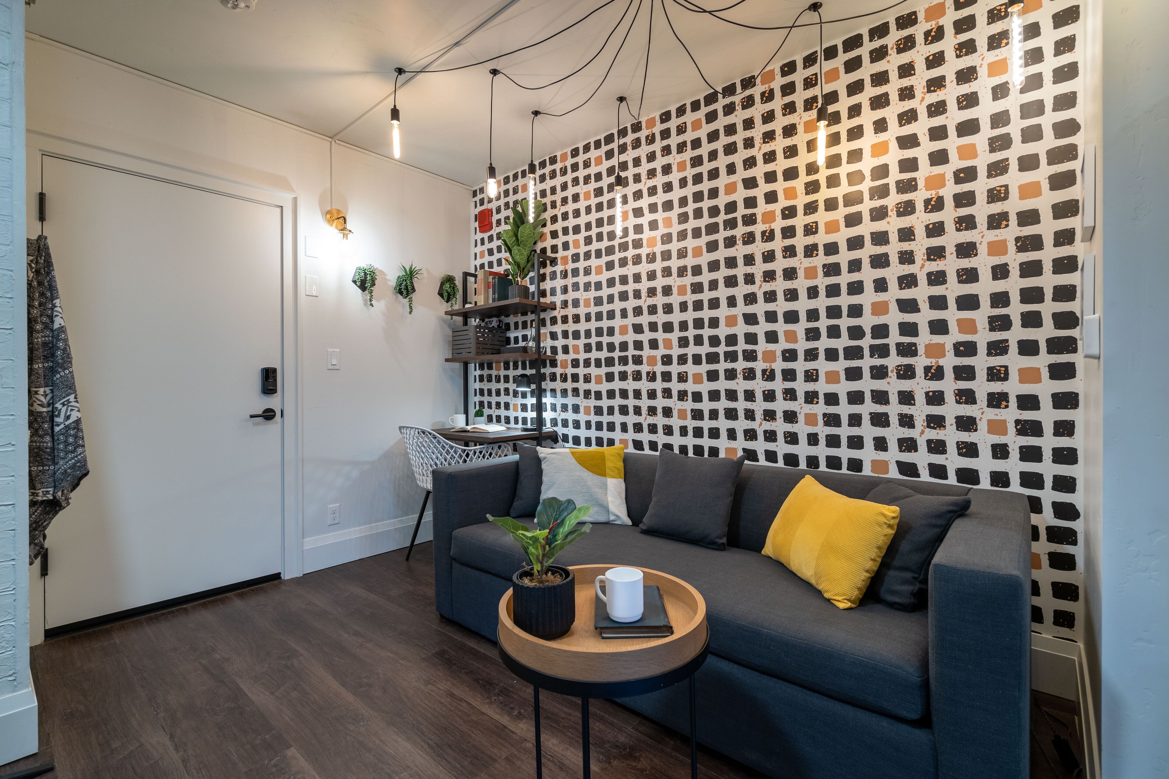 A modern living room with a grey sofa, a small wooden table with a plant and a mug, a patterned wall, and a white door.