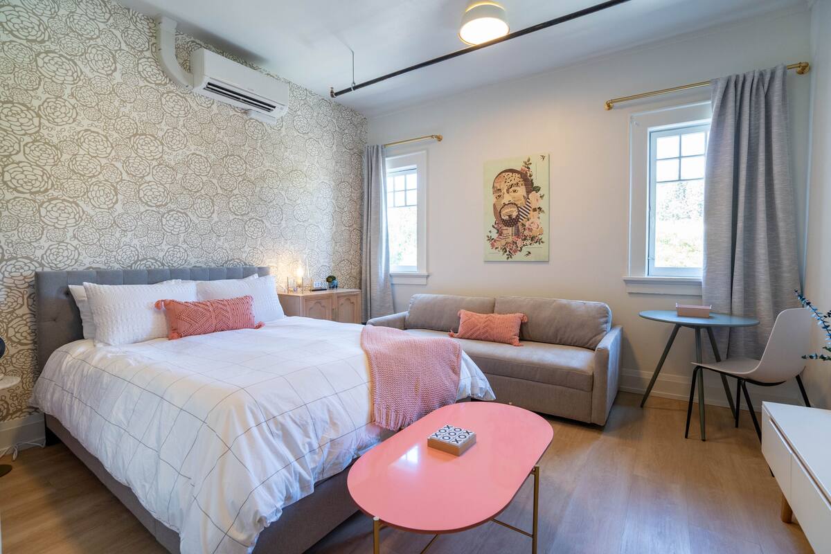A bedroom with a bed, a sofa, a small table, and a chair. There is a window with curtains, a wall-mounted air conditioner, and a wall with a floral pattern.