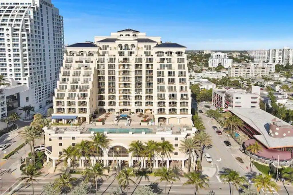 A multi-story hotel with a central courtyard, surrounded by palm trees and adjacent to a road.