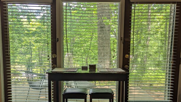A room with a wooden floor, a table with two stools, and a view through large windows with blinds.