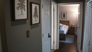 A hallway with framed tree artwork on the walls, a bed with a wooden headboard in the background, and a nightstand with a lamp.