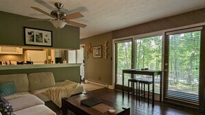 A living room with a sofa, coffee table, and a dining area with a table and chairs. There is a ceiling fan, a kitchen area with a microwave and refrigerator, and large windows with blinds.