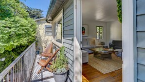 Een balkon met houten stoelen en een bloempot, een woonkamer met een bank en salontafel, en uitzicht op een boom.