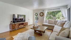 Een moderne woonkamer met een flatscreen televisie, een houten tv-meubel, een beige bank en een groot raam met gordijnen.