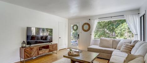A modern living room with a flat-screen TV, wooden entertainment unit, beige sofa, and a large window with curtains.