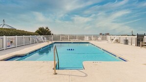 Une piscine rectangulaire entourée d’une terrasse de sable, avec une clôture blanche et des chaises longues.