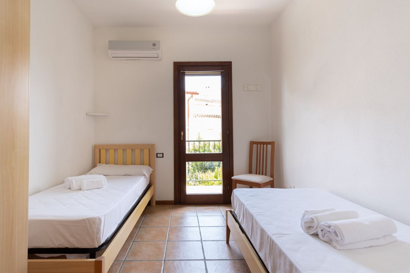 A small, clean room with two single beds, a wooden chair, a small table, and a door leading to a balcony.