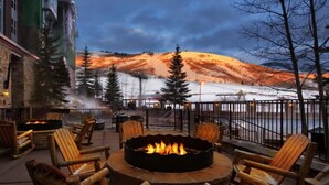 Un espace extérieur aménagé avec des sièges, comprenant un foyer entouré de chaises en bois, offrant une vue sur un paysage enneigé avec une montagne et un bâtiment en arrière-plan.