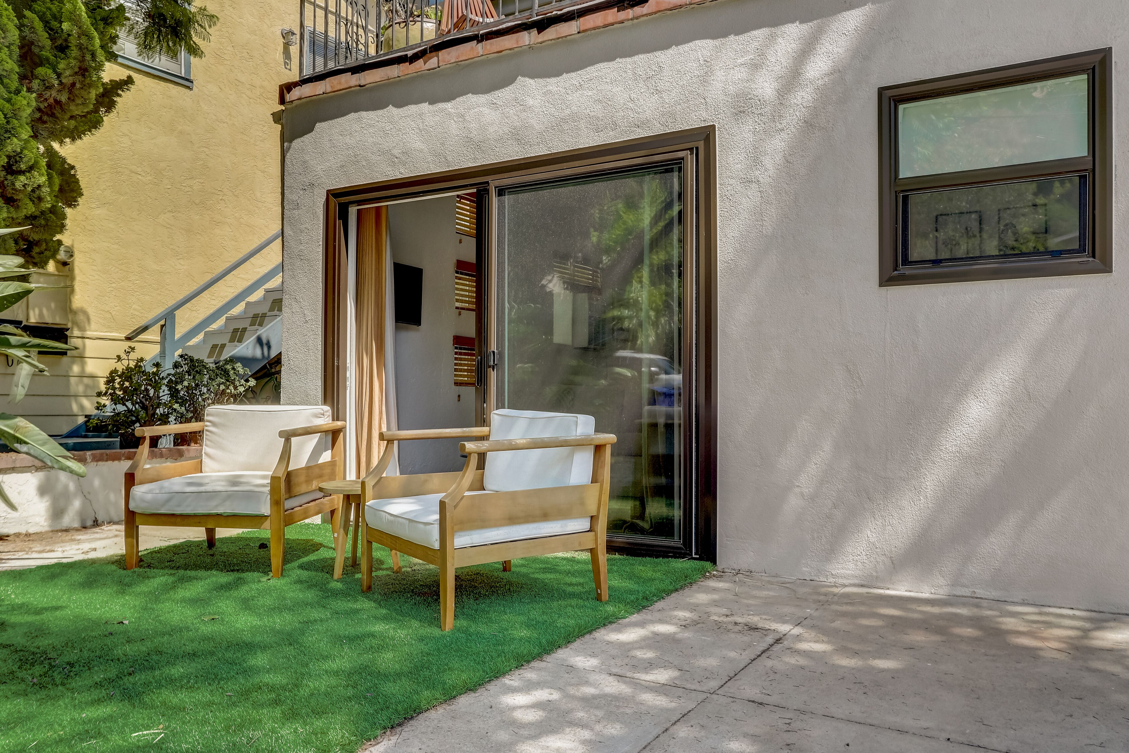 Un edificio moderno con una porta scorrevole in vetro, un piccolo patio con due sedie di legno e una scala che conduce a un livello superiore.