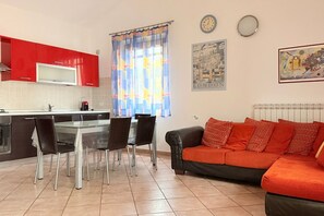 Une cuisine avec des meubles de rangement rouges, une table à manger avec des chaises, un canapé, une fenêtre avec des rideaux et une horloge au mur.
