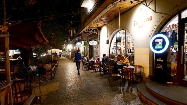A nighttime street scene with outdoor seating at a restaurant, a wine bar, and a shop with a circular sign.
