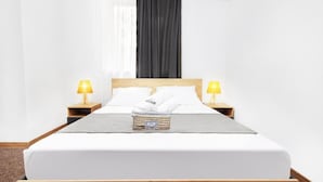A neatly made bed with white linens, two bedside lamps, a dark curtain, and a small wicker basket on the bed.