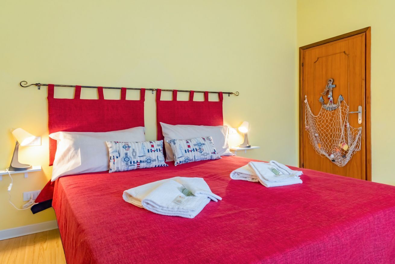 Cama bem arrumada com roupa de cama vermelha, almofadas brancas e cabeceira vermelha. Há duas luminárias de mesa, uma porta de madeira com detalhe em rede e um candeeiro fixado na parede.