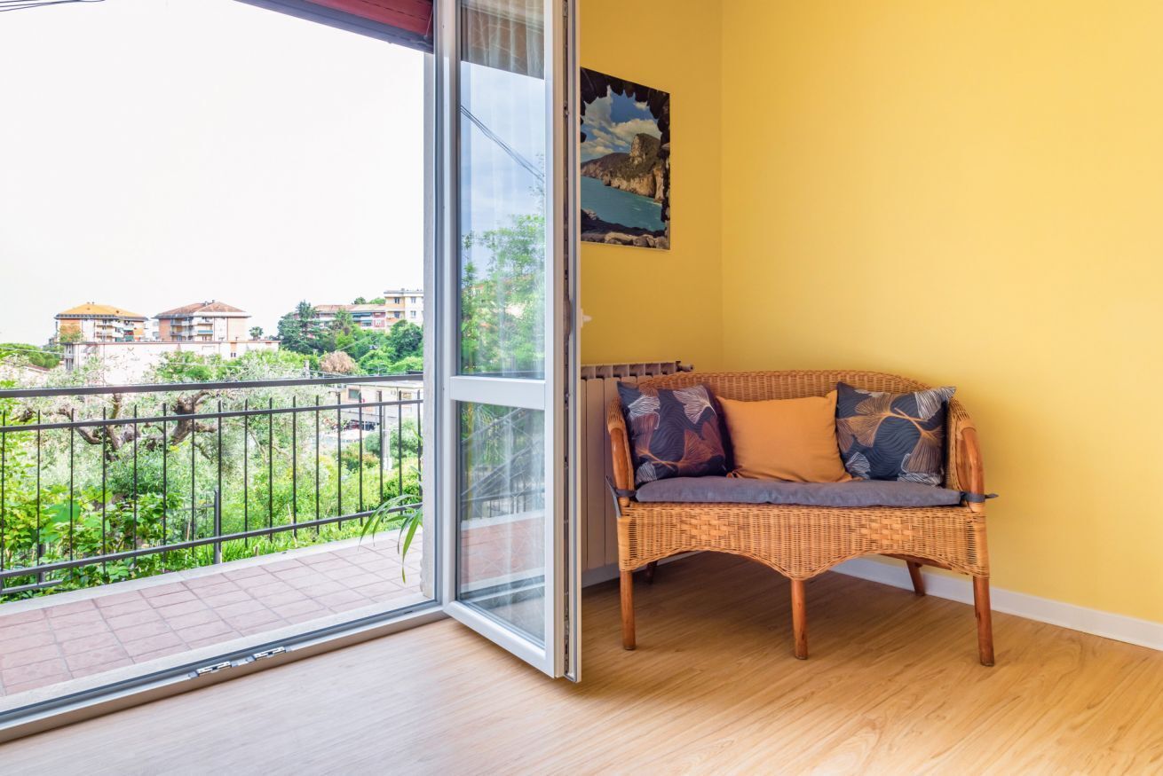 Een balkon met een rieten bank, een glazen deur en een gele muur met een schilderij.