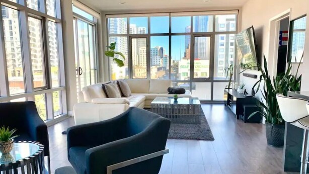 Una sala de estar moderna con sofá, sillón y mesa de centro, ventanas amplias con vista a la ciudad y un área de comedor con una planta.