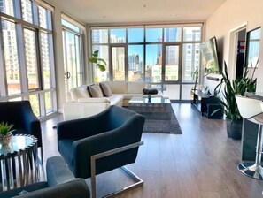 A modern living room with a sofa, armchair, and coffee table, large windows with a city view, and a dining area with a plant. - Downtown Luxury - 2 bed / 2 bath / 2 parking (San Diego)