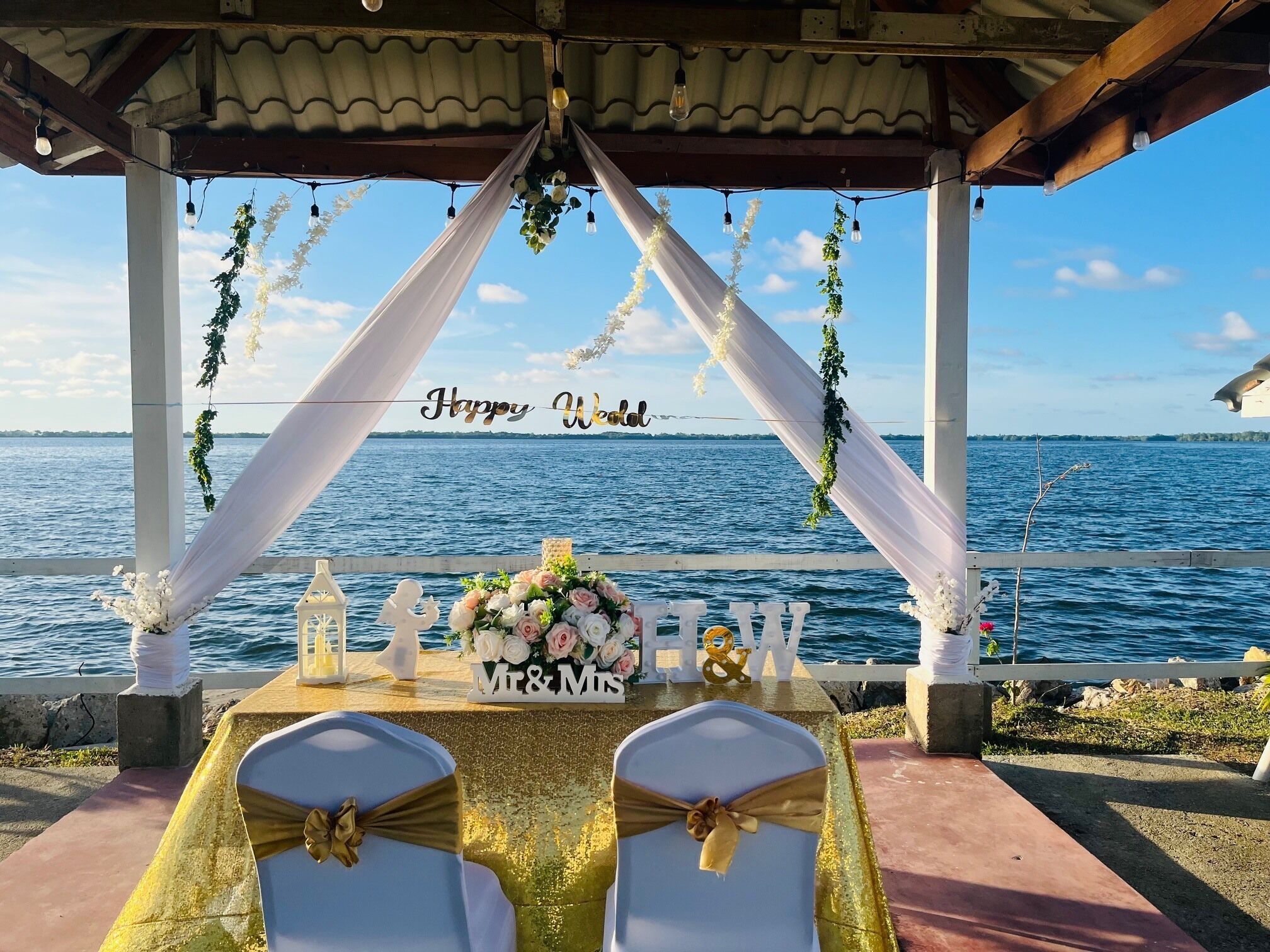A wedding setup with a view of the sea, featuring a table with 'Mr & Mrs' sign, chairs with gold sashes, and a 'Happy Wedding' banner.