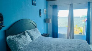 Un dormitorio con cama, aire acondicionado, mesita de noche, lámpara y vista al mar a través de cortinas.