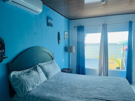 Un dormitorio con cama, aire acondicionado, mesita de noche, lámpara y vista al mar a través de cortinas.