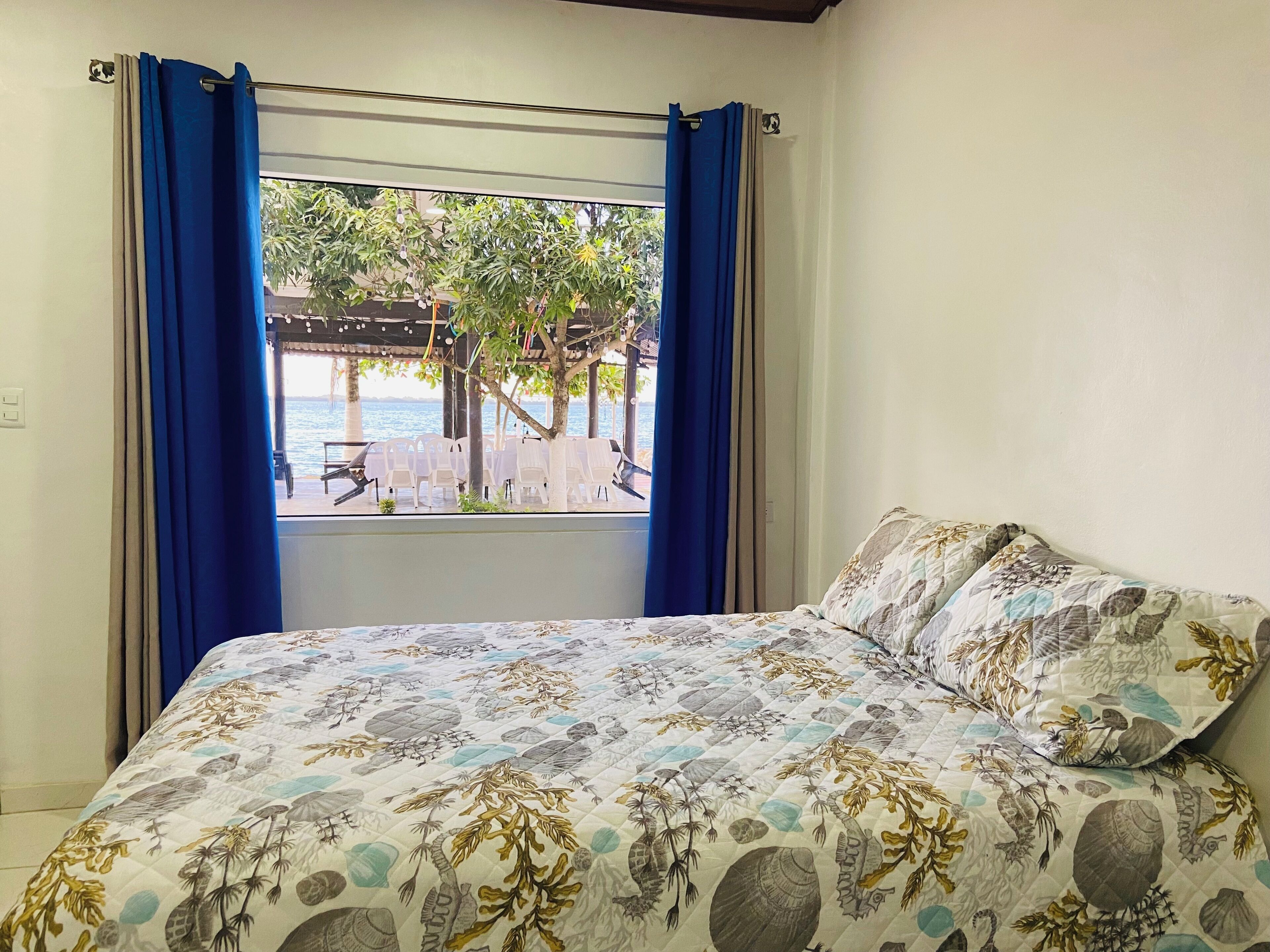Un dormitorio con una cama, un ventanal con cortinas azules y vista a un área exterior con sillas y una mesa.