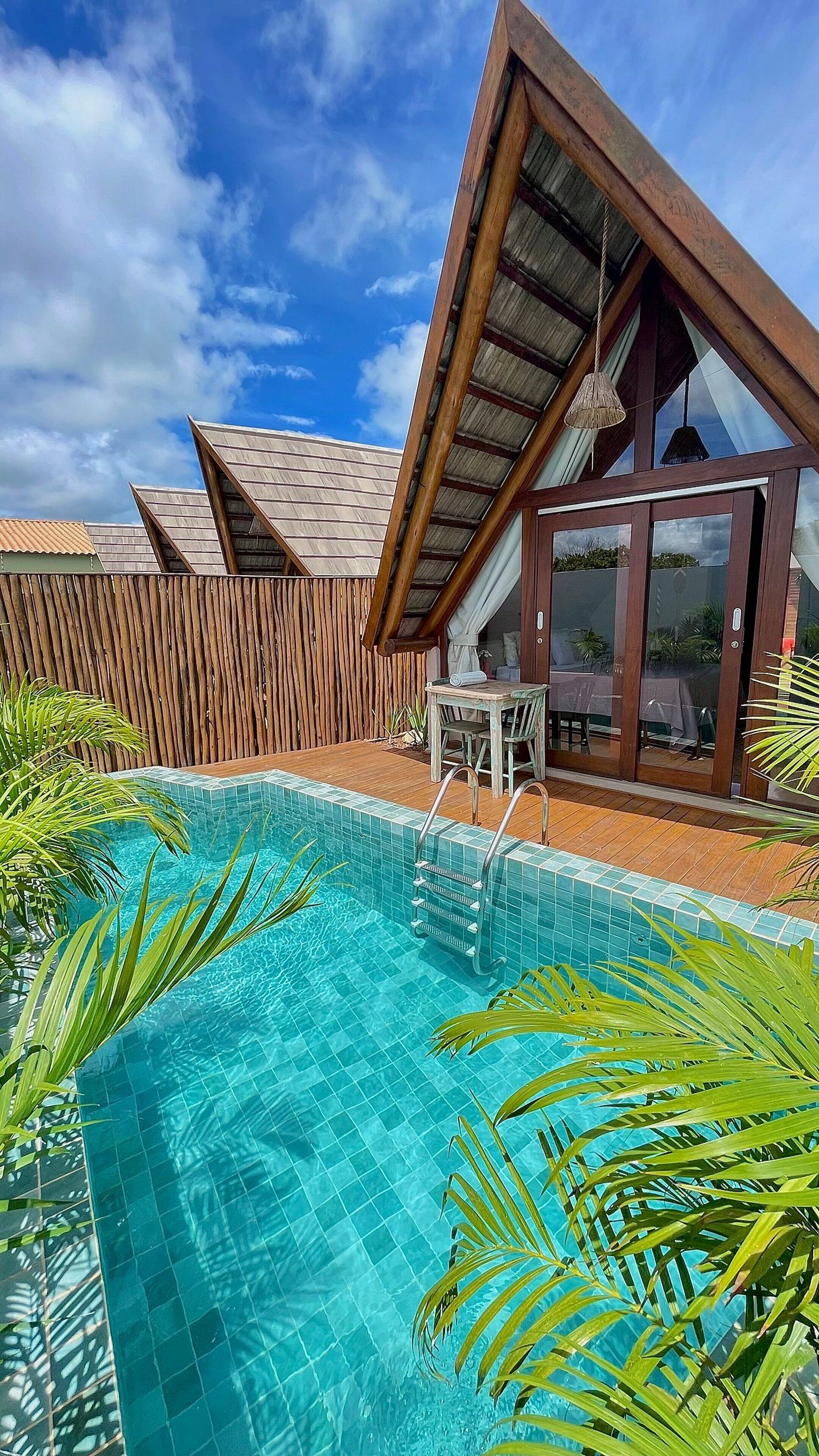 Un área junto a la piscina con cubierta de madera, un espacio de estar con paredes de vidrio y una piscina azul cristalina.