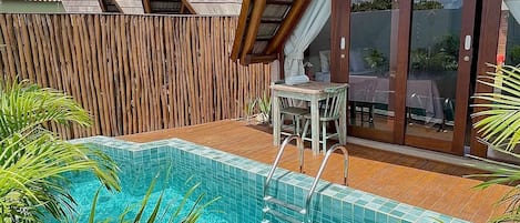 Un área junto a la piscina con cubierta de madera, un espacio de estar con paredes de vidrio y una piscina azul cristalina.