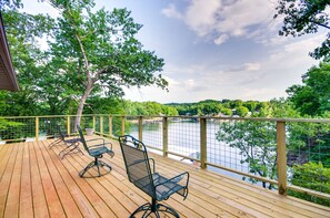 Une terrasse en bois avec des chaises en métal, donnant sur un lac, entourée d’arbres et agrémentée d’une rambarde en bois.
