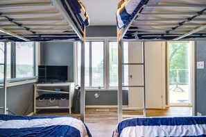 Ett studentrum med våningssängar, ett skrivbord med en dator och en tv. Det finns fönster med persienner, en dörr som leder ut och ett litet förvaringsskåp med väskor.