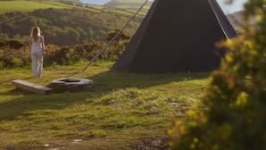 A person standing near a black teepee on a grassy hill with a view of the sea in the background. - Black Shanti Tipi 2 With Ocean View (Tintagel)