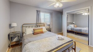 Een slaapkamer met een bed, nachtkastje, lamp, plafondventilator, raam met gordijnen en een spiegel.