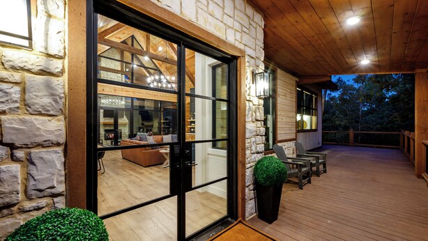 Une terrasse en bois avec des sièges d’extérieur, un mur en pierre et une porte vitrée donnant sur un espace intérieur doté d’une cheminée et d’un coin salon.