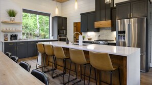 Une cuisine moderne dotée d’un grand îlot central, de tabourets de bar, de meubles de couleur sombre, d’appareils électroménagers en acier inoxydable et de luminaires suspendus.