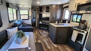 A compact RV interior with a seating area, kitchenette, and a large window providing a view of the outdoors. - Country RV Glamping with lake, mountain and orchard views! (Chelan)