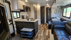 A compact RV kitchen with a microwave, refrigerator, and a sink. There's a small dining area with a table and a couch. - Country RV Glamping with lake, mountain and orchard views! (Chelan)
