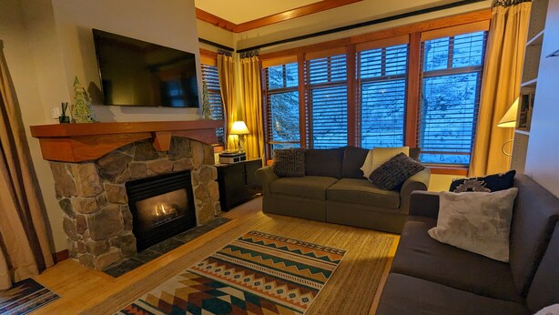 A cozy living room with a stone fireplace, a sofa, and a large window with blinds. - Beautiful End-Unit with Private Balcony and hot tub + pool. (Solitude)