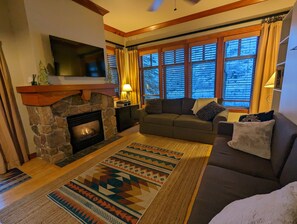 A cozy living room with a stone fireplace, a sofa, and a large window with blinds. - Beautiful End-Unit with Private Balcony and hot tub + pool. (Solitude)
