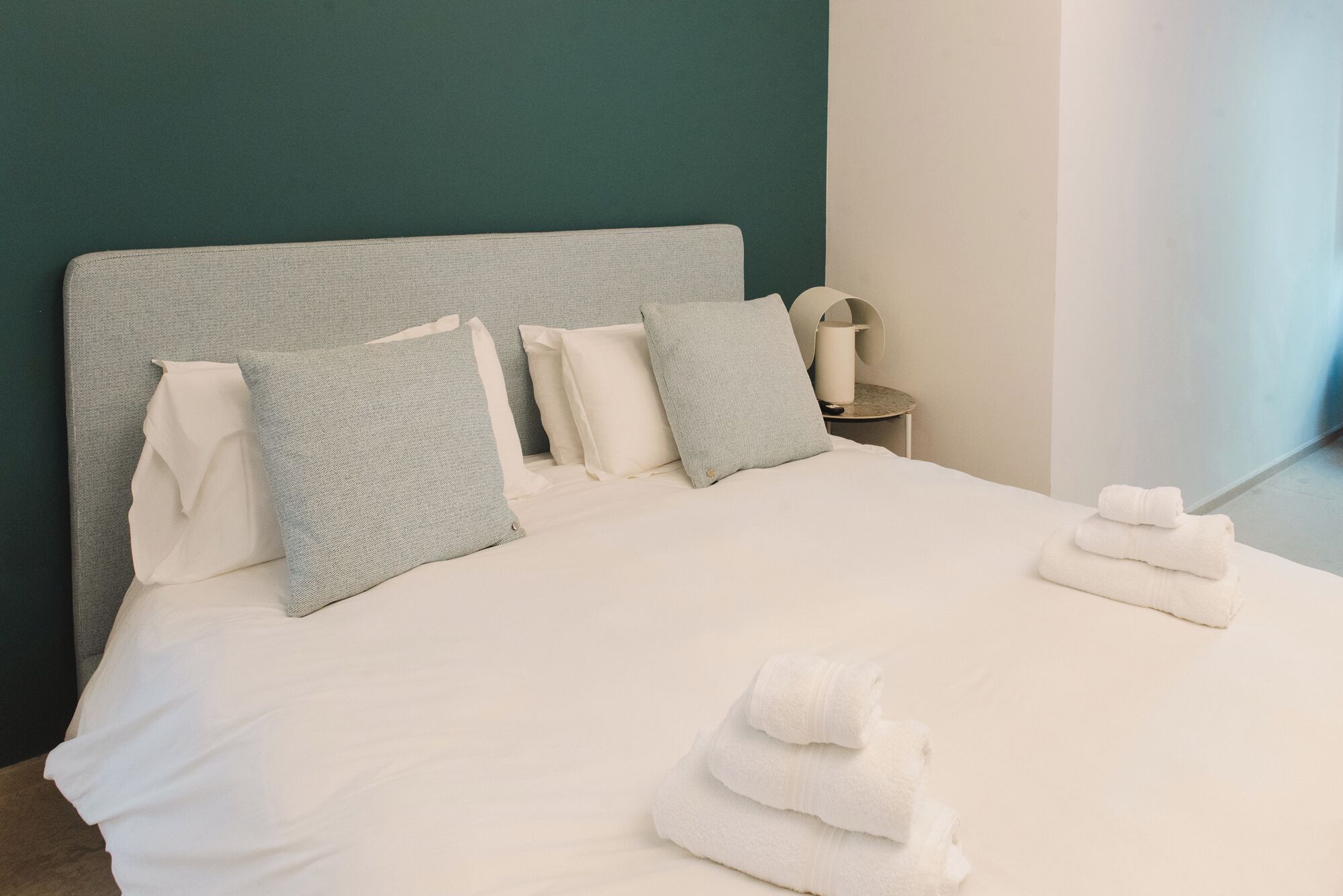 A neatly made bed with pillows, a bedside table with a lamp, and a towel stack.
