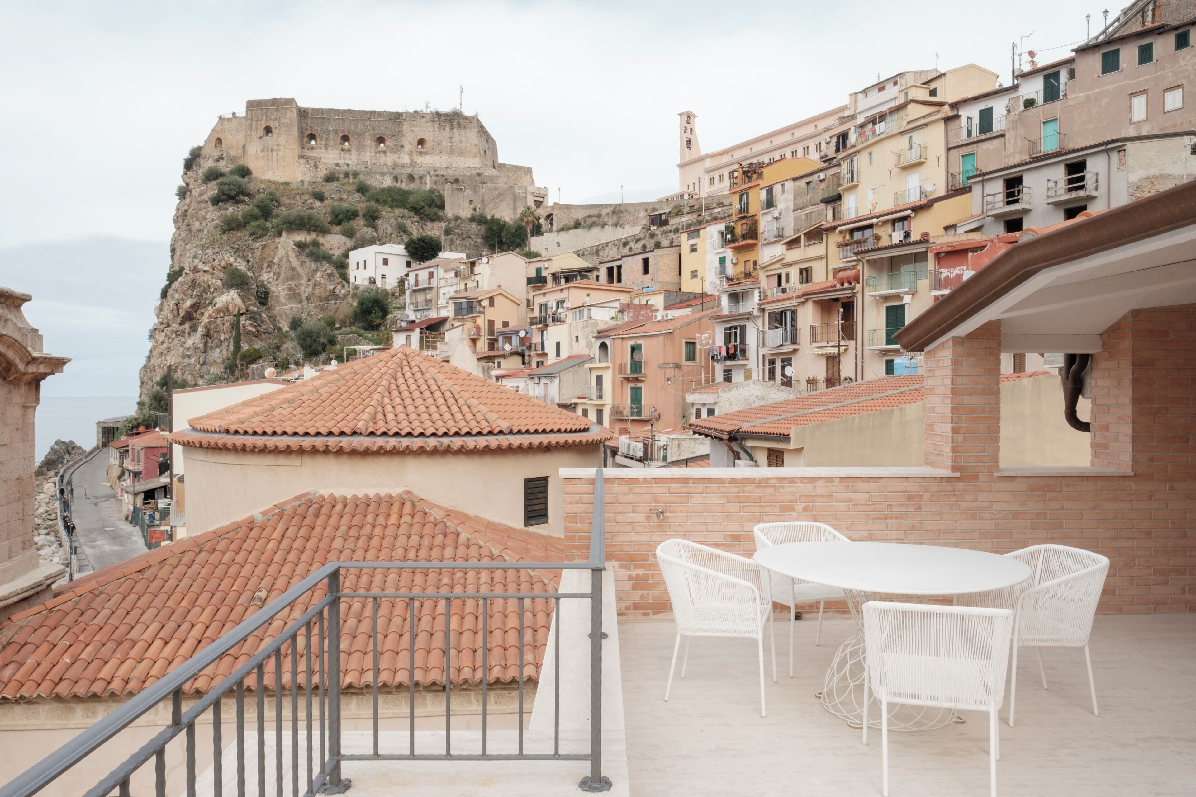 Una terrazza con tavolo e sedie, vista su un'antica fortezza e una cittadina costiera con edifici dalle facciate in terracotta.