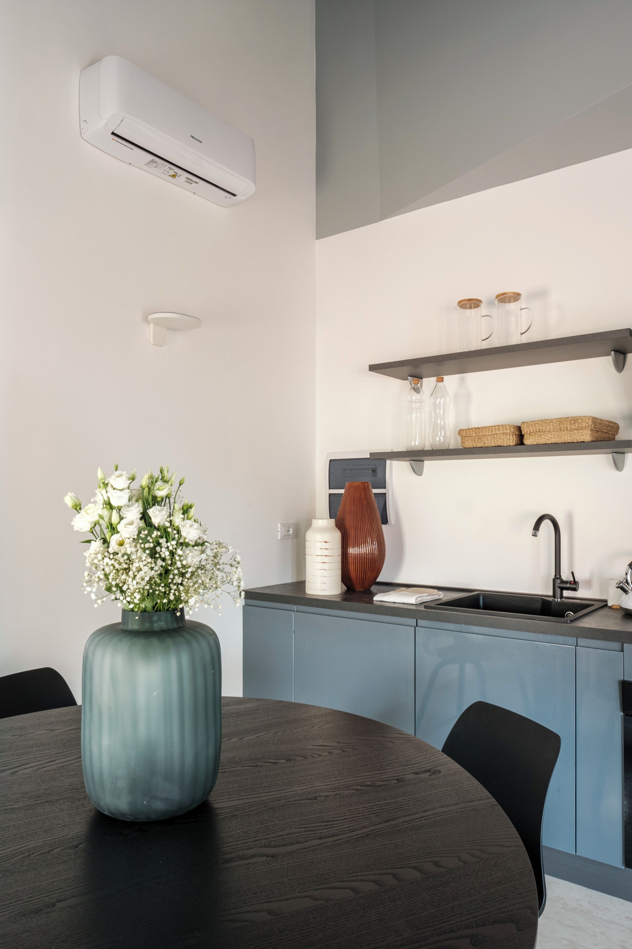 Une cuisine moderne équipée d’un climatiseur intégré, une table à manger ronde ornée d’un vase de fleurs et une chaise noire.